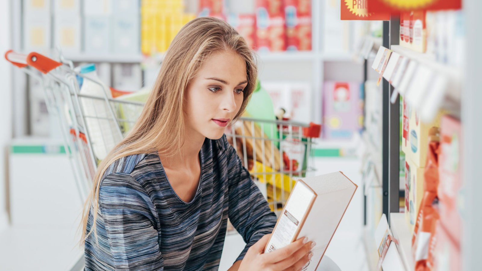 Young woman doing grocery shopping at the supermarket and reading a food label with ingredients on a box, using Nutrigenomics to inform health choices